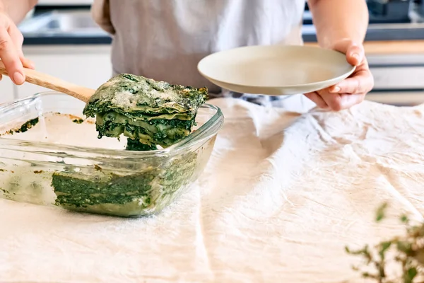 Woman\'s hands putting one porzione of homemade lasagna with spinach and ricotta in a plate from glass baking dish. Traditional italian cuisine. Selective focus.