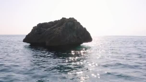Big rocky stone in wavy sea water. Sun reflection on blue water ripples. — Stock Video