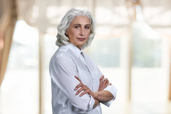Portrait of confident senior woman with crossed arms standing indoors. — Stock Photo, Image