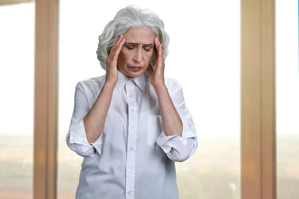 Mulher idosa sobrecarregada que sofre de dor de cabeça. Janela do escritório no fundo. — Fotografia de Stock