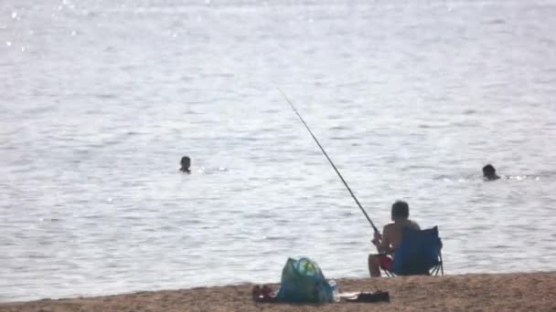 Uomo seduto in riva al mare a pescare. Persone che nuotano in mare. — Video Stock