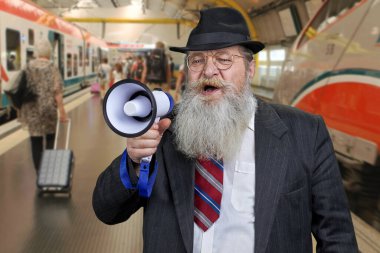 Kendine güvenen sakallı adam megafona konuşuyor. Bulanık tren istasyonu arka planı.