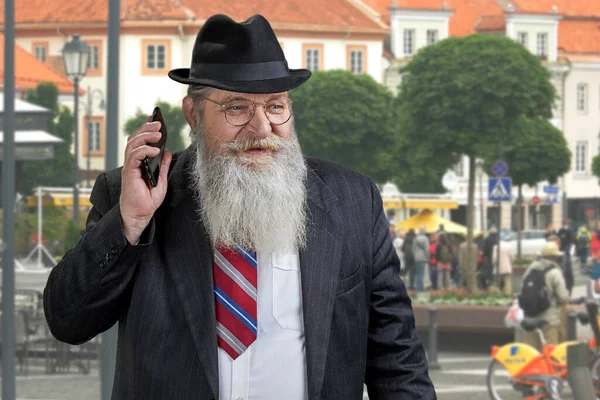 Senior skäggig affärsman i hatt ringer ett samtal och tittar bort stående utomhus. — Stockfoto
