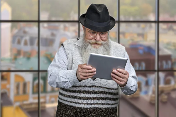 Stary człowiek kaukaski za pomocą tabletu cyfrowego na tle okna z widokiem na budynki miejskie. — Zdjęcie stockowe