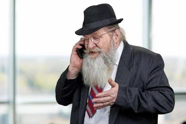 Stary biznesmen negocjuje przez telefon komórkowy stojący na niewyraźnym tle okna biurowego. — Zdjęcie stockowe