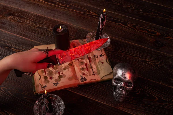 Close-up female hand holds knife over the satanic bible and candles. — Stock Photo, Image