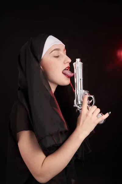 Jonge non zuster houdt zilveren revolver omhoog en likt het. — Stockfoto