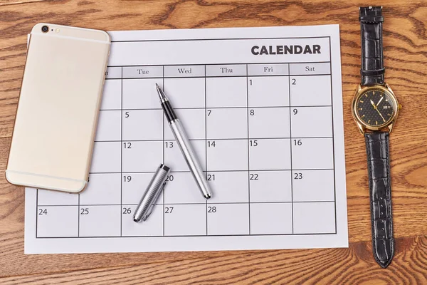 Calendar with smartphone pen and watch clock on brown wooden desk. Business appointment accessories top view flat lay.