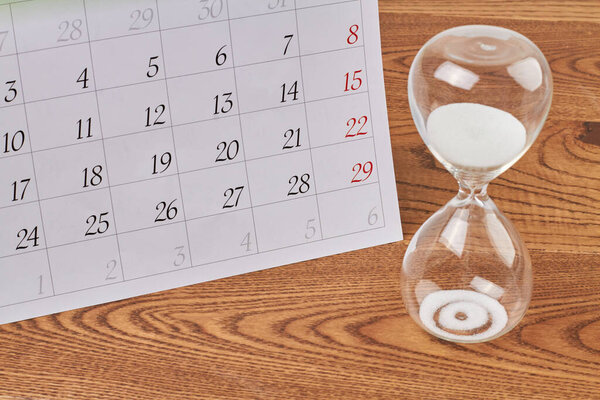 Close-up hourglass with white sand and calendar on wooden desk. Countdown sandglass timer.