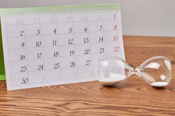 Hourglass with white sand and month calendar on wooden desk. Sandglass countdown timer and calendar.