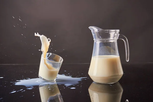 Spilling glass milk in motion and glass jug on reflective desk. Messy milk splashes on reflective mirror surface.