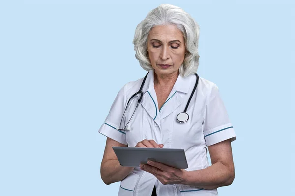 Mature female doctor using digital tablet on light blue background. Senior confident woman doctor holding computer tablet.