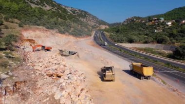 Yol kenarındaki dağ ocağında çalışan ağır makinelerin hava aracı görüntüsü. Madencilik endüstrisi, maden taşımacılığı.