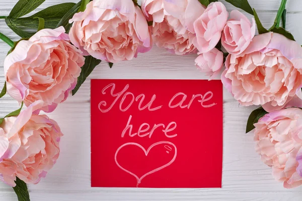 You are here handwriting on red card. Peonies on white desk.