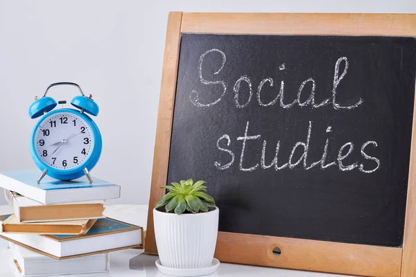 Still life blackboard with social studies words. Houseplant with stack of books and alarm clock.