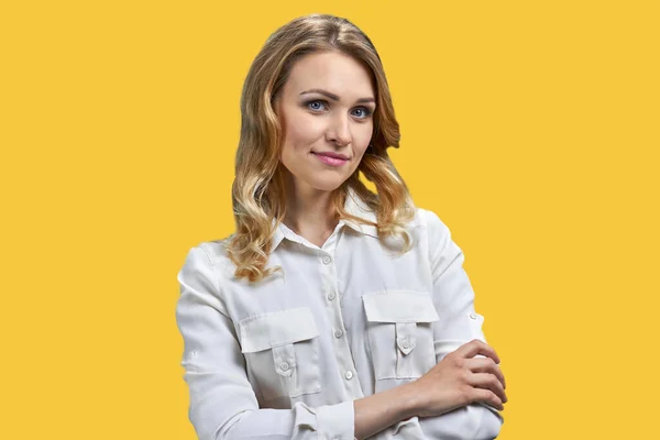 Beautiful young woman in white blouse keeping arms folded while standing on yellow background. Woman fashion model looking at camera.