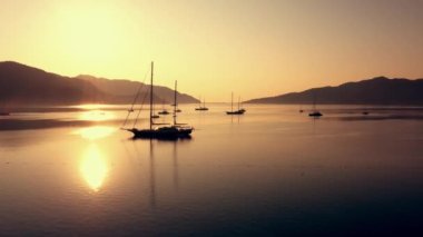 View of calm sea bay with parked boats during sunset. Mountains and orange evening sky in the background. Tranquil summer scene.