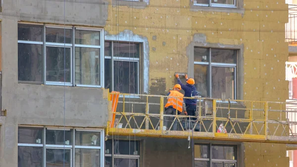 Workers are insulating the wall of a building. Construction site with working men on the elevator. panels. Hanging suspended cradle elevator.