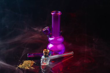 Composition of smoking accessories with pipe and pile of weed. Purple glass bong still life.