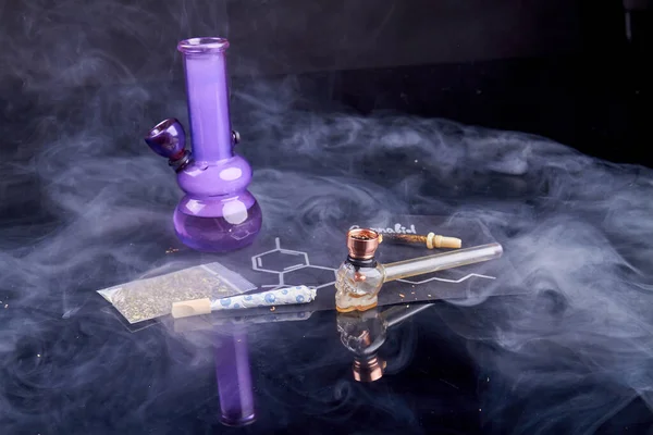 Weed smoking devices and lots of smoke on black background. Bong with pipe and joint.