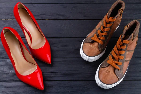 Red female high heel shoes and brown male sneakers. Top view mens and womens shoes on dark wooden background.