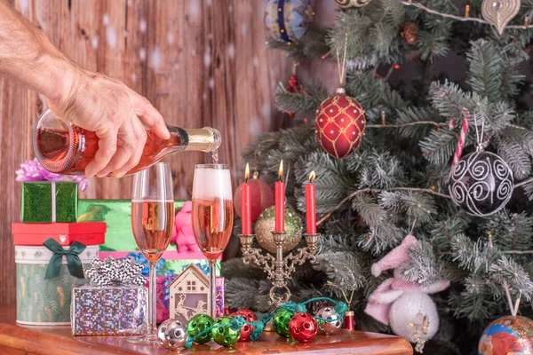 男人们的手把香槟酒从瓶子里倒入香槟酒中，在一棵新年树的背景下，献上礼物. — 图库照片