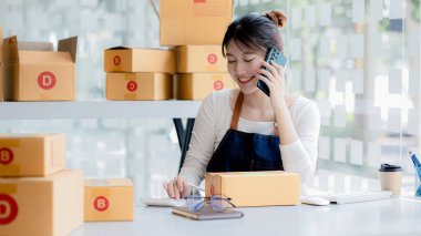 Asyalı bir kadın siparişi doğrulamak için bir müşteriyle telefonda konuşuyor, bir online mağazası var, özel bir nakliye şirketi aracılığıyla paketleyip gönderiyor. Çevrimiçi satış ve online alışveriş kavramları.