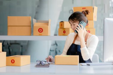 Asyalı bir kadın siparişi doğrulamak için bir müşteriyle telefonda konuşuyor, bir online mağazası var, özel bir nakliye şirketi aracılığıyla paketleyip gönderiyor. Çevrimiçi satış ve online alışveriş kavramları.