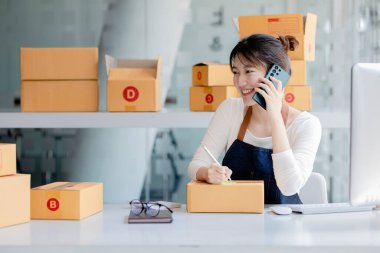 Asyalı bir kadın siparişi doğrulamak için bir müşteriyle telefonda konuşuyor, bir online mağazası var, özel bir nakliye şirketi aracılığıyla paketleyip gönderiyor. Çevrimiçi satış ve online alışveriş kavramları.