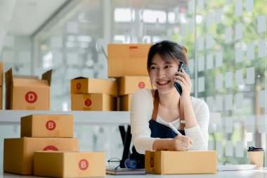 Asyalı bir kadın siparişi doğrulamak için bir müşteriyle telefonda konuşuyor, bir online mağazası var, özel bir nakliye şirketi aracılığıyla paketleyip gönderiyor. Çevrimiçi satış ve online alışveriş kavramları.