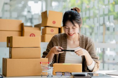Paket kutularının önünde fotoğraf çekmek için akıllı telefon kullanan bir kadın. Malları paketlemek için paket kutuları, özel kurye şirketleri aracılığıyla mal teslimatı. Çevrimiçi satış ve online alışveriş kavramları.