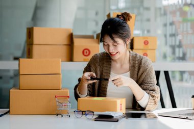 Paket kutularının önünde fotoğraf çekmek için akıllı telefon kullanan bir kadın. Malları paketlemek için paket kutuları, özel kurye şirketleri aracılığıyla mal teslimatı. Çevrimiçi satış ve online alışveriş kavramları.