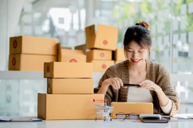 Paket kutularının önünde fotoğraf çekmek için akıllı telefon kullanan bir kadın. Malları paketlemek için paket kutuları, özel kurye şirketleri aracılığıyla mal teslimatı. Çevrimiçi satış ve online alışveriş kavramları.