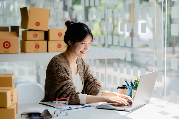 A beautiful Asian business owner opens an online store, she is checking orders from customers via laptop, sending goods through a courier company, concept of a woman opening an online business.