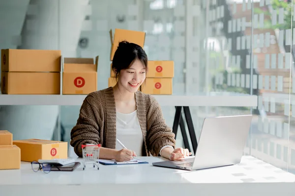 Güzel bir Asyalı işletme sahibi online bir dükkan açıyor, müşterilerin siparişlerini dizüstü bilgisayarla kontrol ediyor, malları bir kurye şirketi aracılığıyla gönderiyor, bir kadının online iş kurma kavramı.