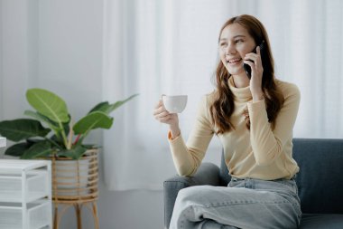 Asyalı bir kadın kahve içiyor ve arkadaşlarıyla telefonda konuşuyor. Oturma odasındaki kanepede. Hafta sonu tatili. Sıkı çalışma..