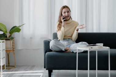 Asyalı bir kadın, oturma odasındaki kanepede arkadaşıyla telefonda sohbet ediyor. Yorucu bir iş gününden sonra hafta sonu molası veriyor. Tatil gevşeme fikirleri.