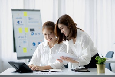 Girişim şirketinin atmosferinde, iki kadın çalışan, satışları artırmak ve yöneticilere raporlar hazırlamak için özetler ve pazarlama planları üzerinde çalışmak üzere fikir alışverişinde bulunuyorlar..