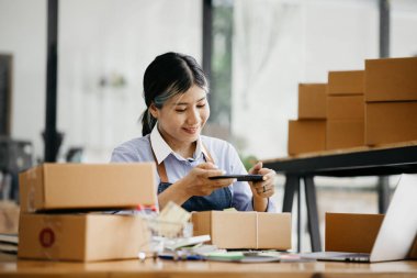 A woman using a smartphone to take pictures in front of parcel boxes, parcel boxes for packing goods, delivering goods through private courier companies. Online selling and online shopping concepts.