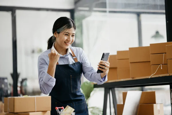 Woman who runs an online store with a happy face is holding her phone to check sales, she sells online and packs goods through a private shipping company. concept of a woman opening an online business