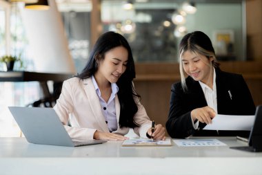 Girişim şirketinin atmosferinde, iki kadın çalışan, satışları artırmak ve yöneticilere raporlar hazırlamak için özetler ve pazarlama planları üzerinde çalışmak üzere fikir alışverişinde bulunuyorlar..