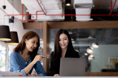 Girişim şirketinin atmosferinde, iki kadın çalışan, satışları artırmak ve yöneticilere raporlar hazırlamak için özetler ve pazarlama planları üzerinde çalışmak üzere fikir alışverişinde bulunuyorlar..