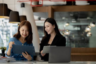 Girişim şirketinin atmosferinde, iki kadın çalışan, satışları artırmak ve yöneticilere raporlar hazırlamak için özetler ve pazarlama planları üzerinde çalışmak üzere fikir alışverişinde bulunuyorlar..