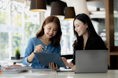 Girişim şirketinin atmosferinde, iki kadın çalışan, satışları artırmak ve yöneticilere raporlar hazırlamak için özetler ve pazarlama planları üzerinde çalışmak üzere fikir alışverişinde bulunuyorlar..