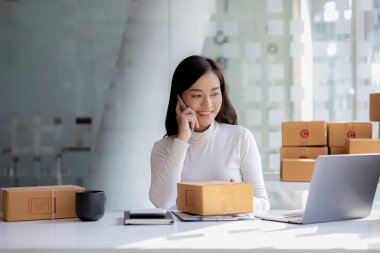 Asyalı bir kadın siparişi doğrulamak için bir müşteriyle telefonda konuşuyor, bir online mağazası var, özel bir nakliye şirketi aracılığıyla paketleyip gönderiyor. Çevrimiçi satış ve online alışveriş kavramları.