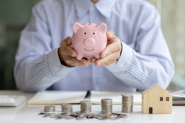 A person is holding a pink pig piggy bank, piles of coins on the table and a house model on it, saving money to spend and managing to buy a future home. Money saving ideas to buy a house or residence.