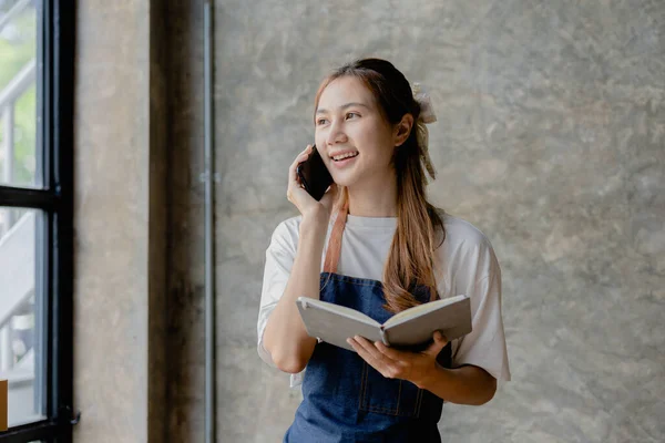 Beautiful Asian business owner opens an online store, She is on the phone with a customer to confirm the order, sending goods through a courier company, concept of a woman opening an online business.