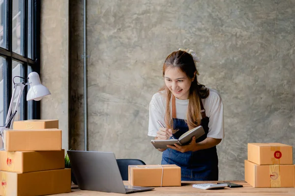 Beautiful Asian business owner opens an online store, She was writing down the order in her notebook, sending goods through a courier company, concept of a woman opening an online business.