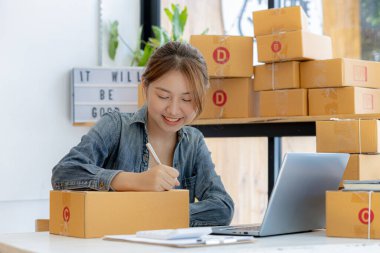Bir e-ticaret şirketini işleten kadın, onlara göndermeden önce müşterilerin bir listesini kağıda yazıyor. Web sitelerinde ve sosyal medyada bir e-ticaret şirketi işletiyor. Çevrimiçi ürün satma kavramı.