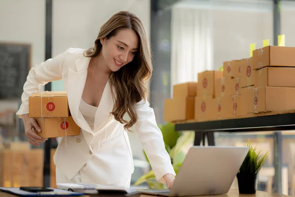 A beautiful Asian business owner opens an online store, she is checking orders from customers via laptop, sending goods through a courier company, concept of a woman opening an online business.